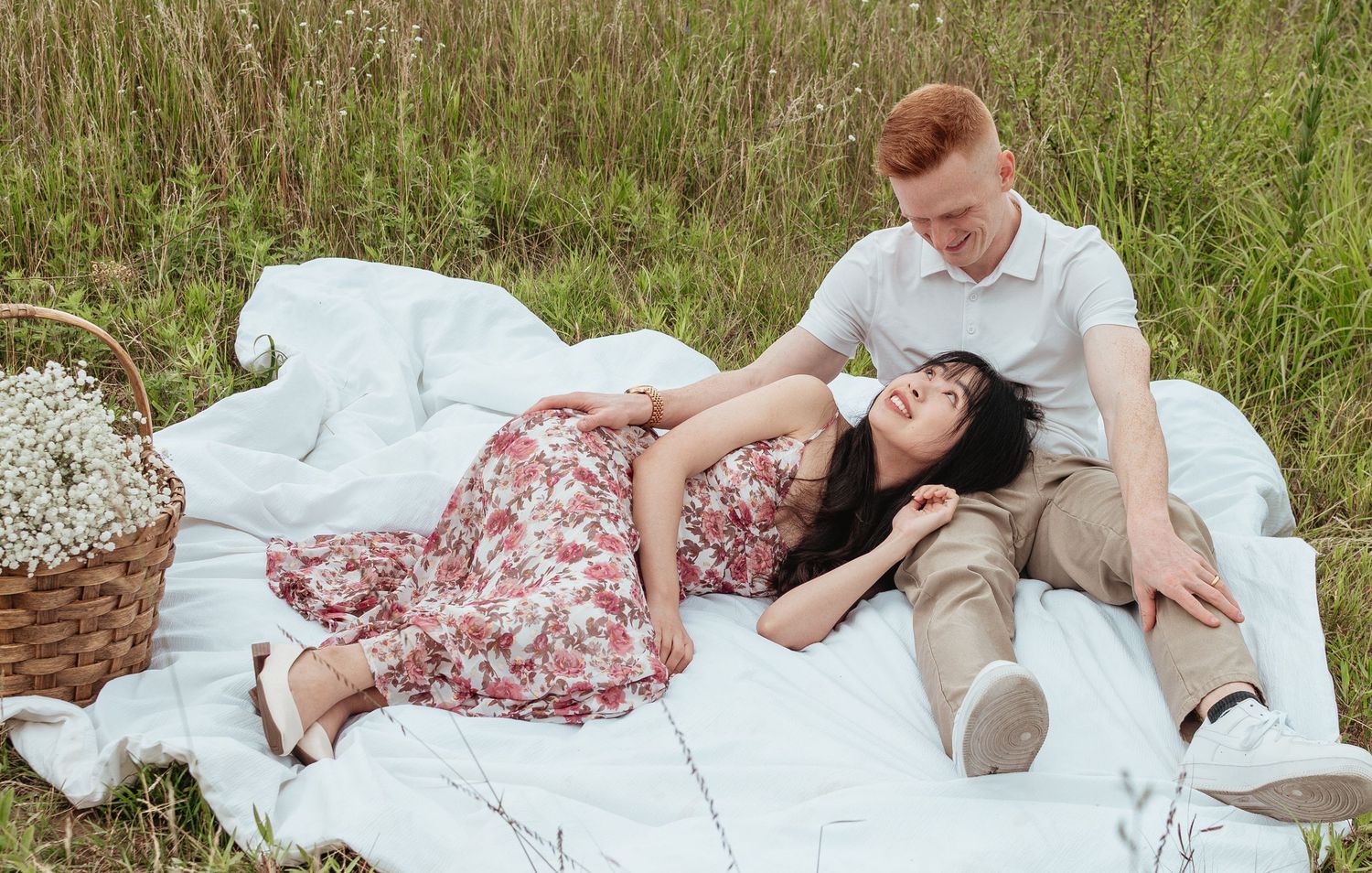A couple lounges on a white blanket in a grassy field during a romantic outdoor picnic.
