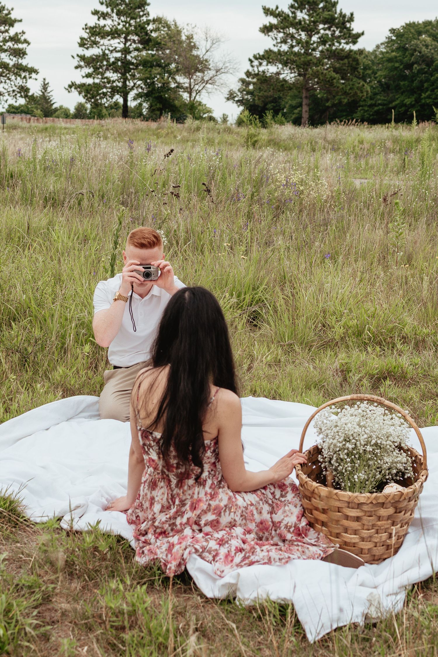 Two people enjoying a romantic picnic on a white blanket in a meadow with a basket of flowers.