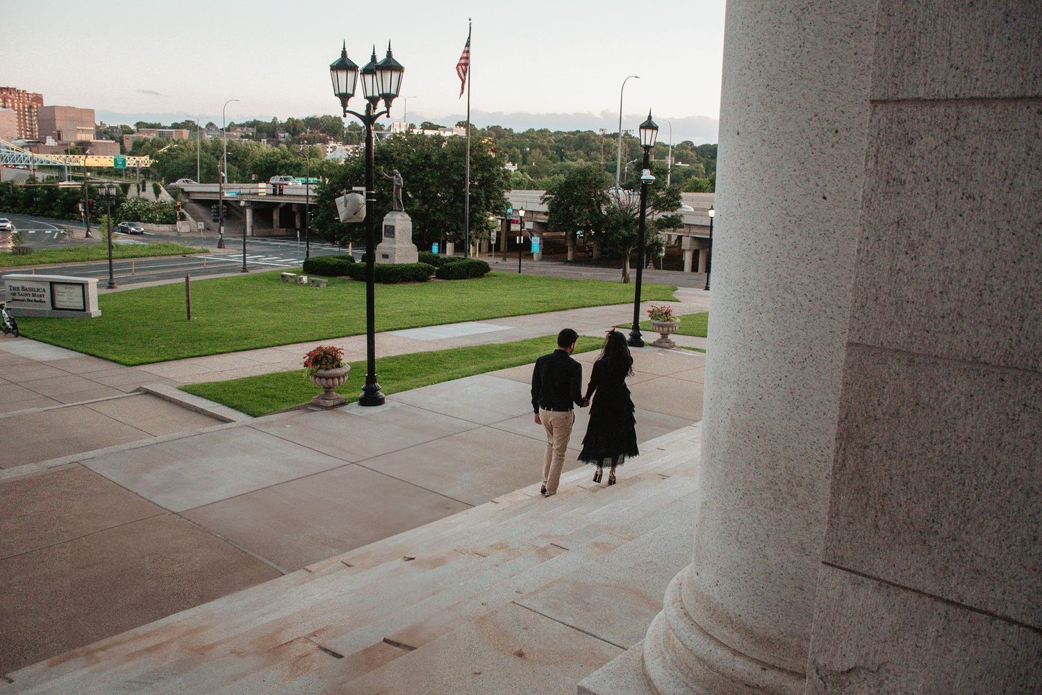 A couple walks down stone steps near classic architecture and lamp posts.