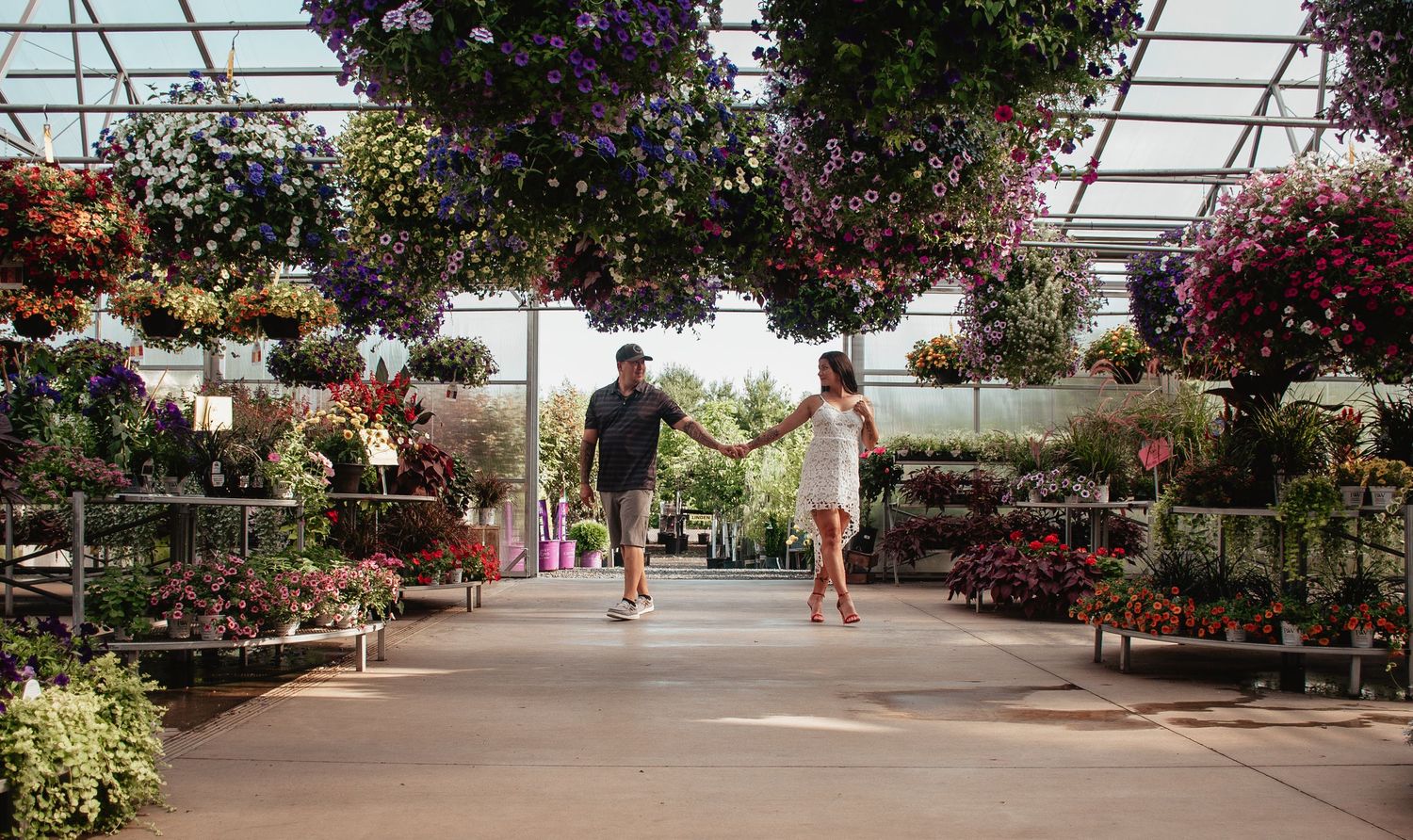 A couple walks through a garden center greenhouse filled with colorful hanging flower baskets.