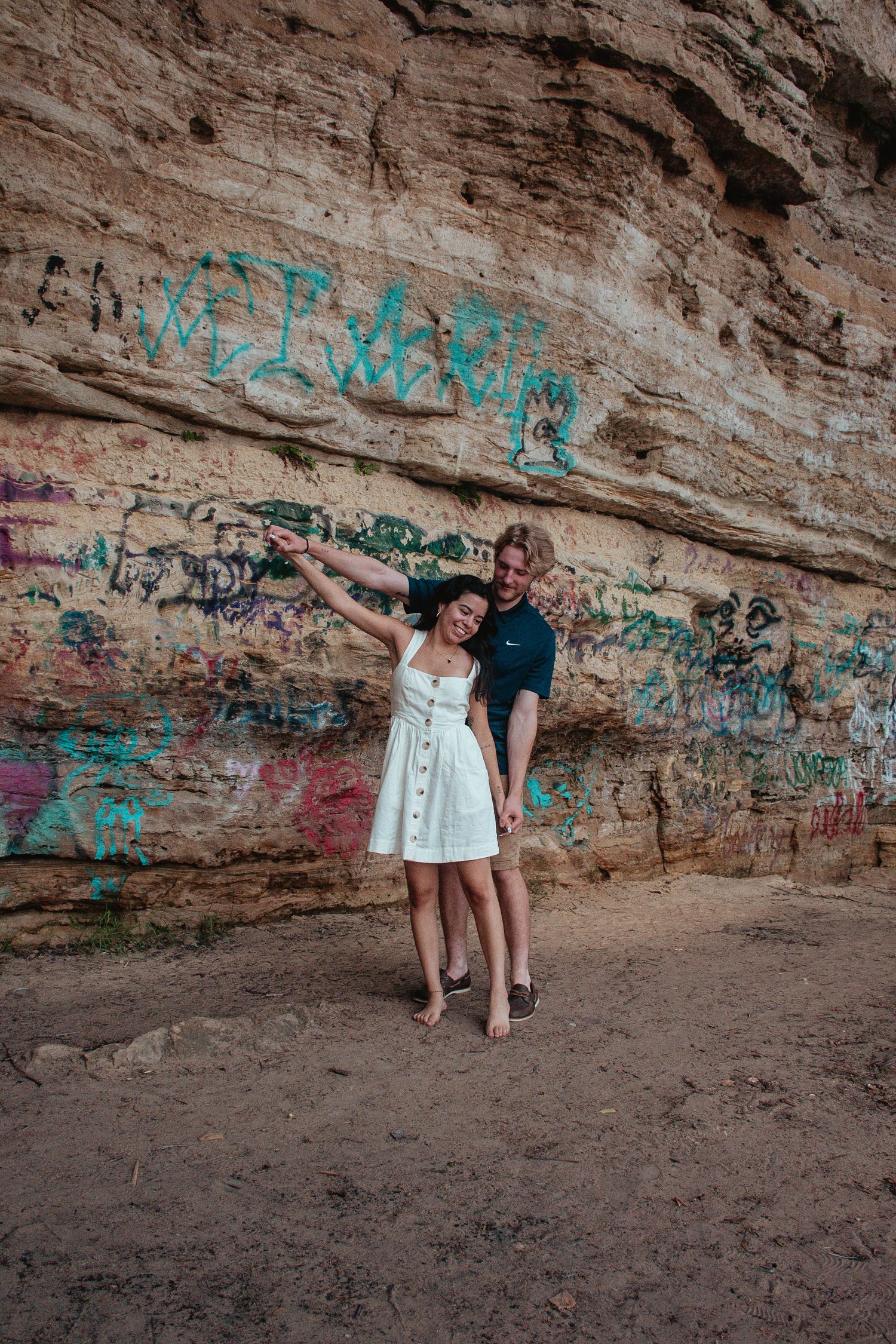 A couple poses playfully against a graffiti-covered rock wall outdoors.
