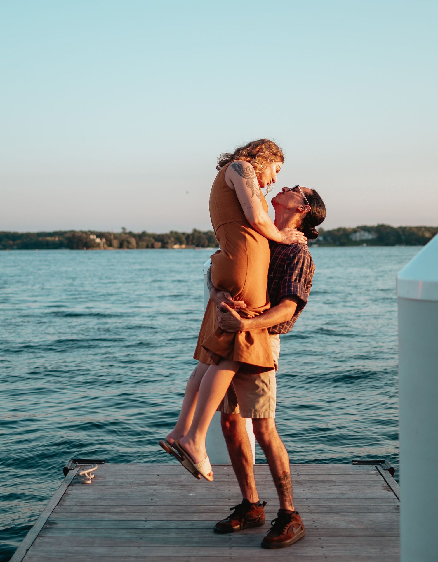 A couple shares an intimate embrace on a wooden dock overlooking a tranquil lake at sunset.