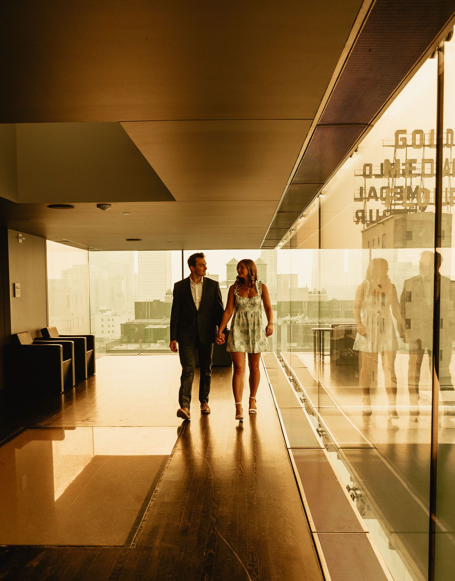 A well-dressed couple walks down a modern hallway with glass walls and warm lighting creating dramatic shadows.