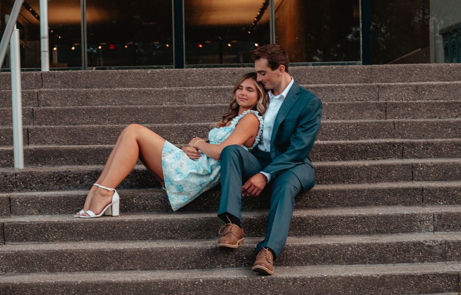A couple sits casually on outdoor concrete steps at sunset in a romantic urban setting.