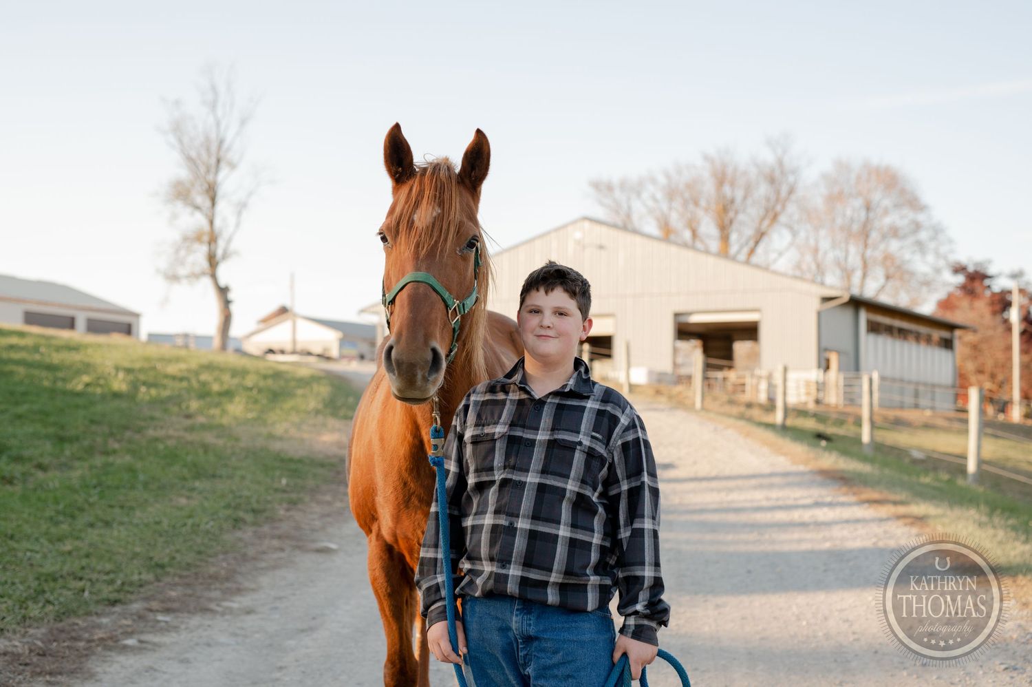 A person in casual attire stands with their chestnut horse on a farm path.