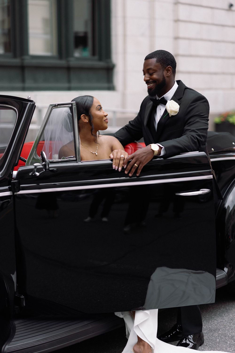 Couple poses with vintage black convertible during urban wedding shoot.