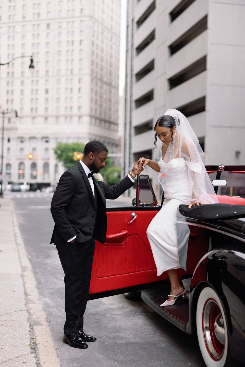 Groom helps bride into classic red vintage car on city street.