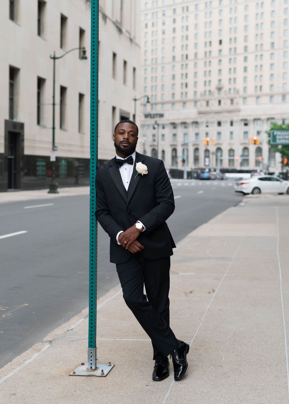 Groom in classic black tuxedo poses against urban building backdrop.