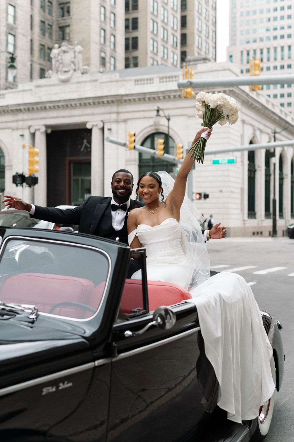 Newlyweds celebrate in vintage black convertible on city street.