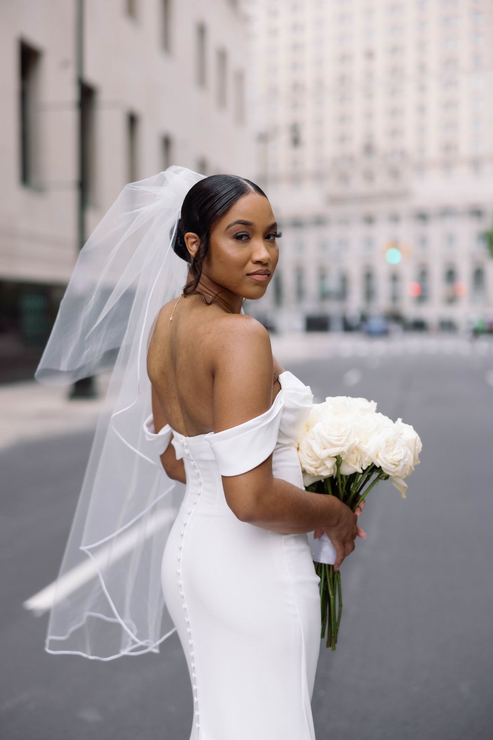 Elegant bride in fitted off-shoulder wedding dress holds white bouquet against city backdrop.
