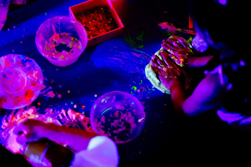 Glowing neon objects and containers under blacklight creating vibrant purple and blue reflections.