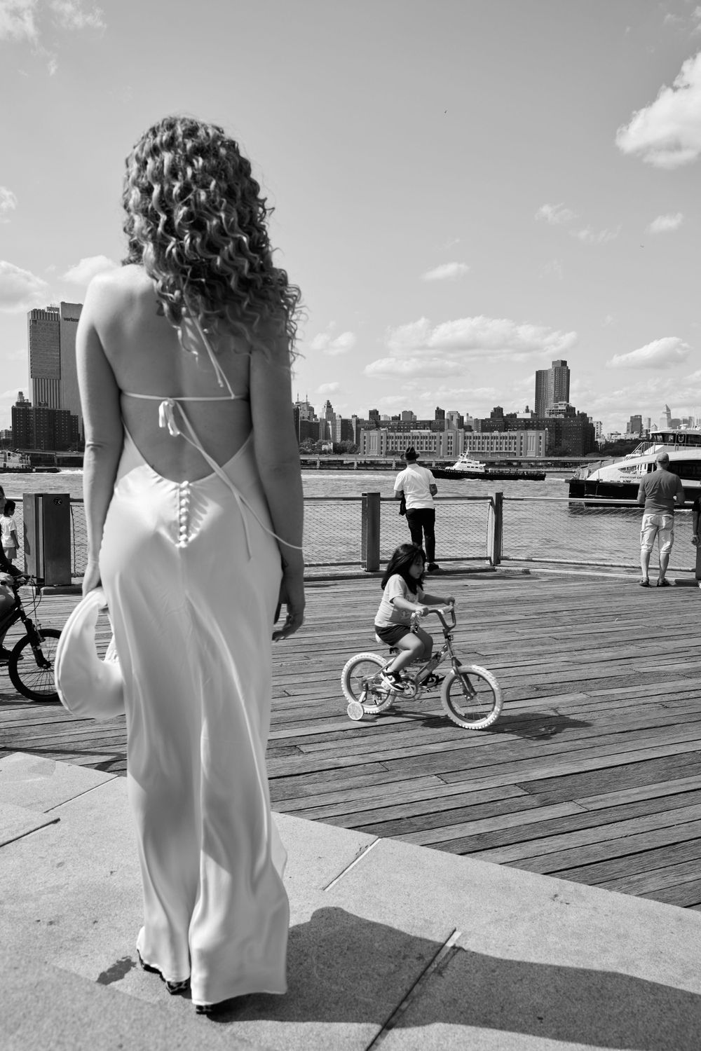 Woman in backless white dress on wooden boardwalk overlooking city waterfront.
