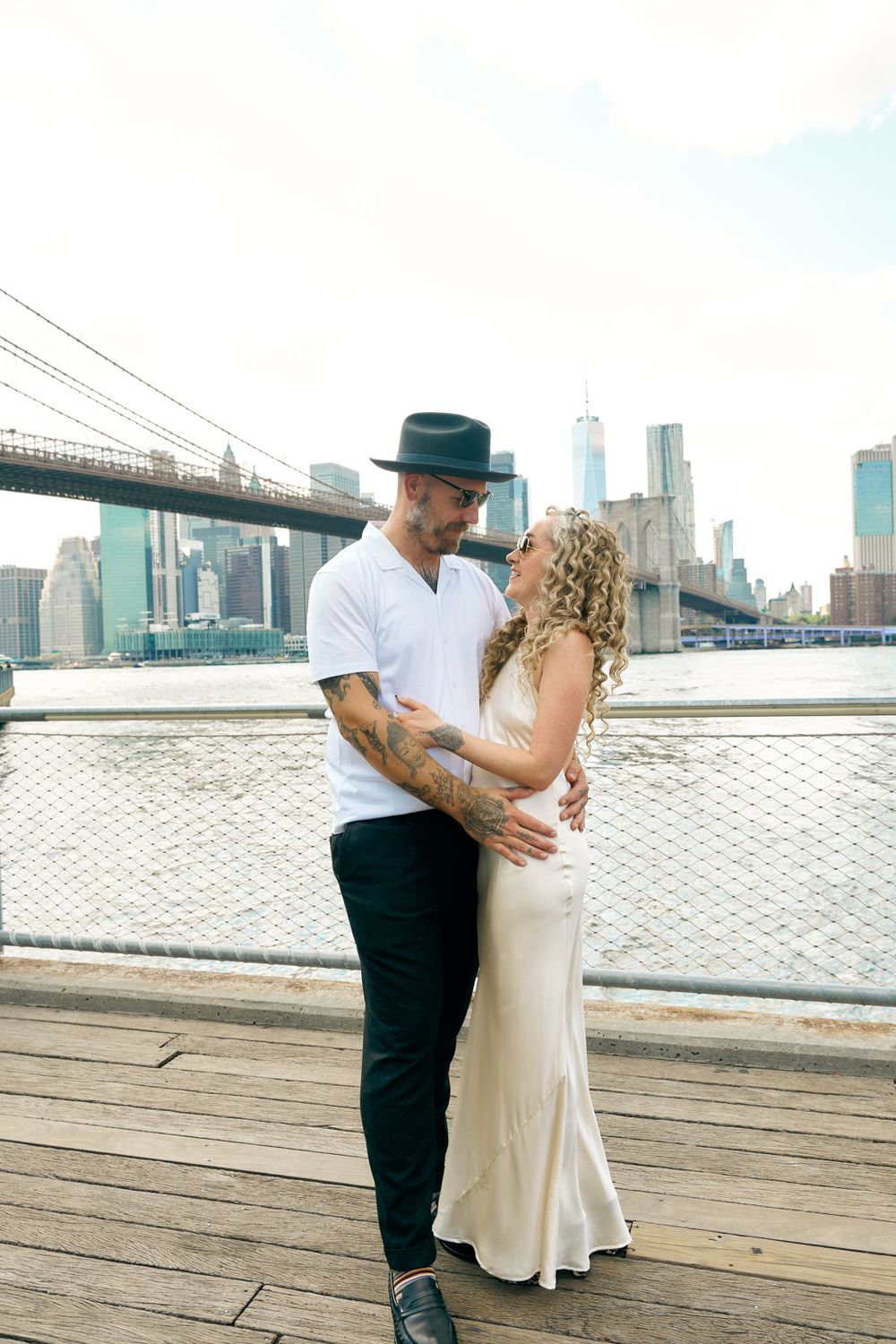 A couple shares an intimate moment on a waterfront deck with the Brooklyn Bridge and NYC skyline in background.