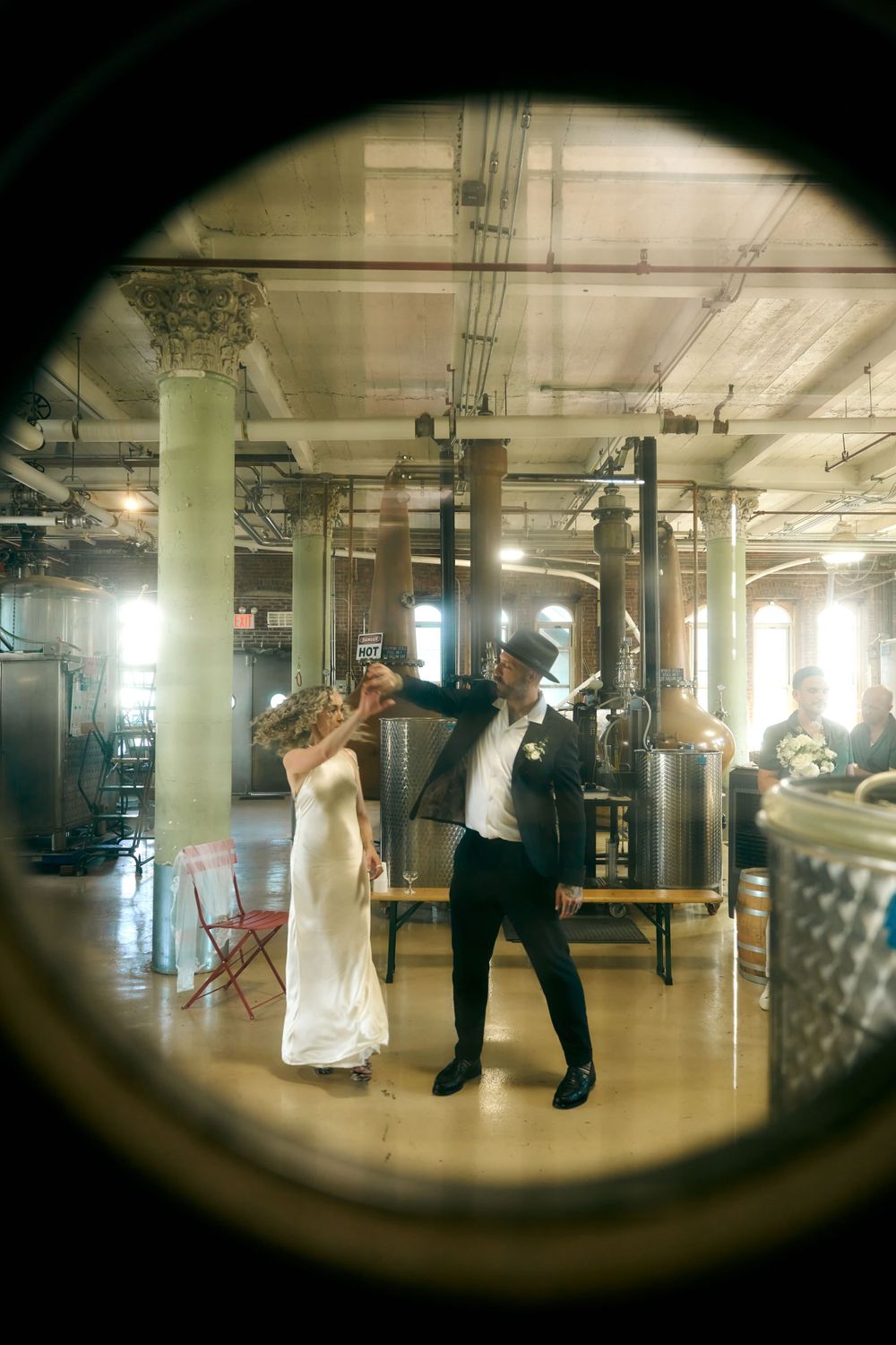 A couple dances in an industrial-style venue viewed through a circular window frame.