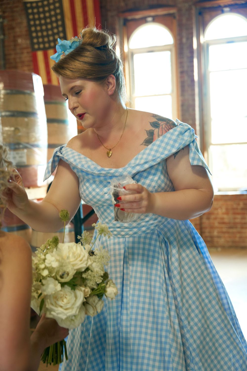 A woman in a blue gingham dress holds flowers in a sunlit brick-walled room.