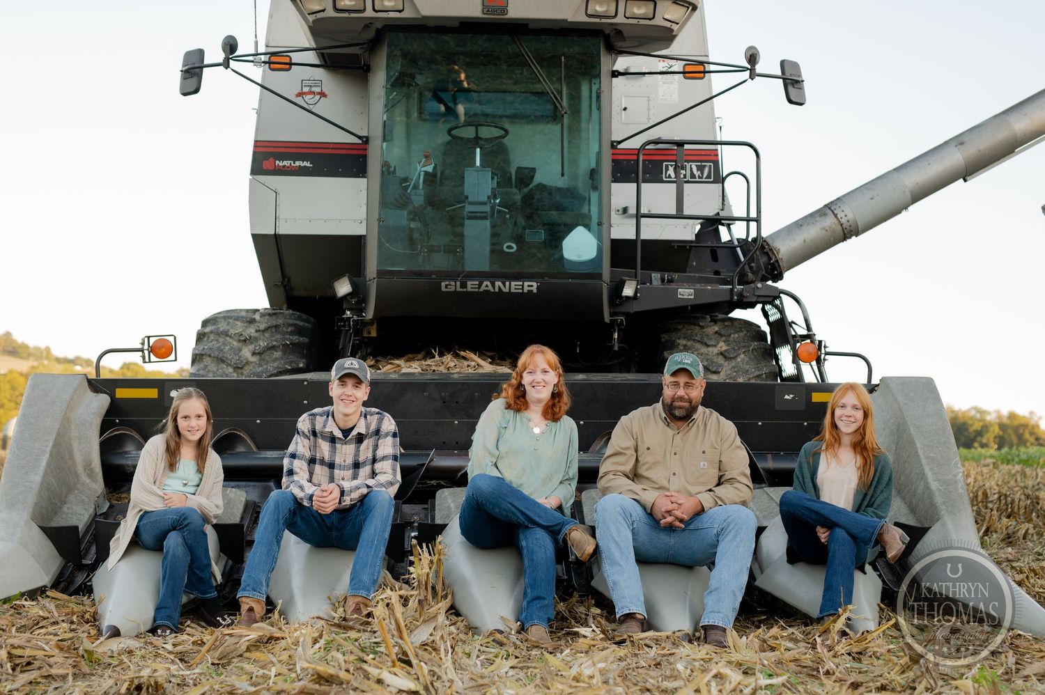 A group sits together in front of a large combine harvester in a farm field.