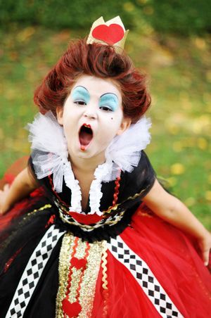 A dramatic Queen of Hearts cosplay costume featuring white makeup, red cape and crown against a blurred green background.