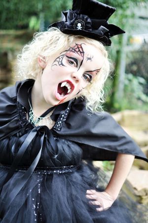 A Halloween costume featuring dramatic vampire makeup, black Victorian-style dress, and mini top hat against an outdoor setting.