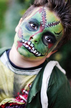 Creative Halloween zombie face paint design featuring green skin, zipper details and gruesome teeth makeup on young person.