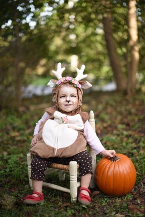 A small deer-costumed child sits in a white chair next to a large orange pumpkin in an autumn woodland setting.