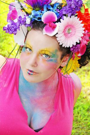 Artistic portrait with vibrant floral crown featuring gerbera daisies and bold fantasy makeup in pink and blue tones.