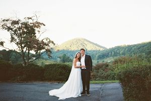 A couple in wedding attire stands on a paved road with scenic mountains and trees in the background.