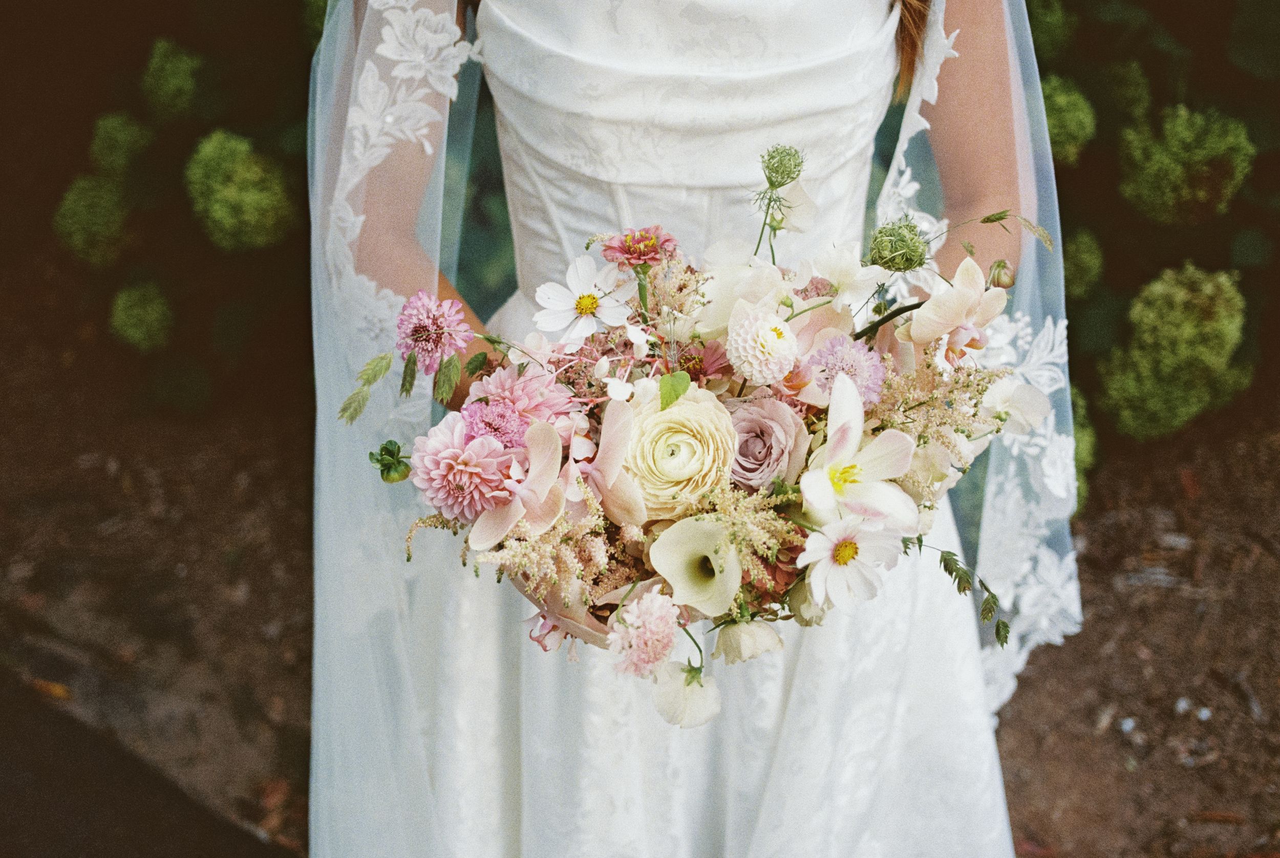 Wedding bouquet composed of delicate pink roses and white florals with lace trimmed dress in the background.