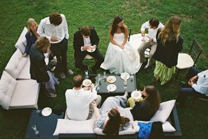 Wedding guests gather for outdoor reception on grass lawn with bistro chairs and white benches around tables.