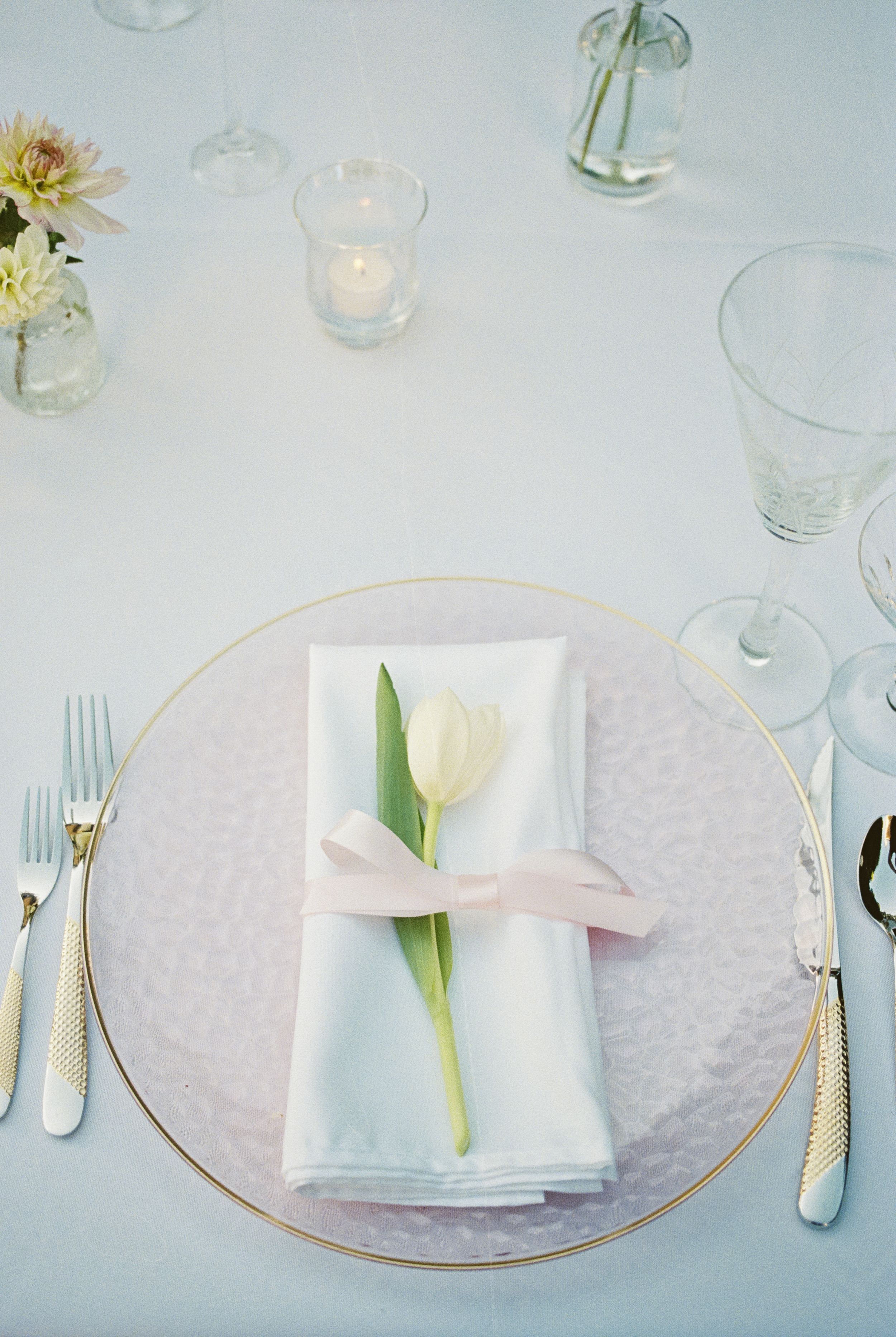 Elegant table setting with white tulip on napkin, gold charger plate and soft candlelight on white tablecloth.