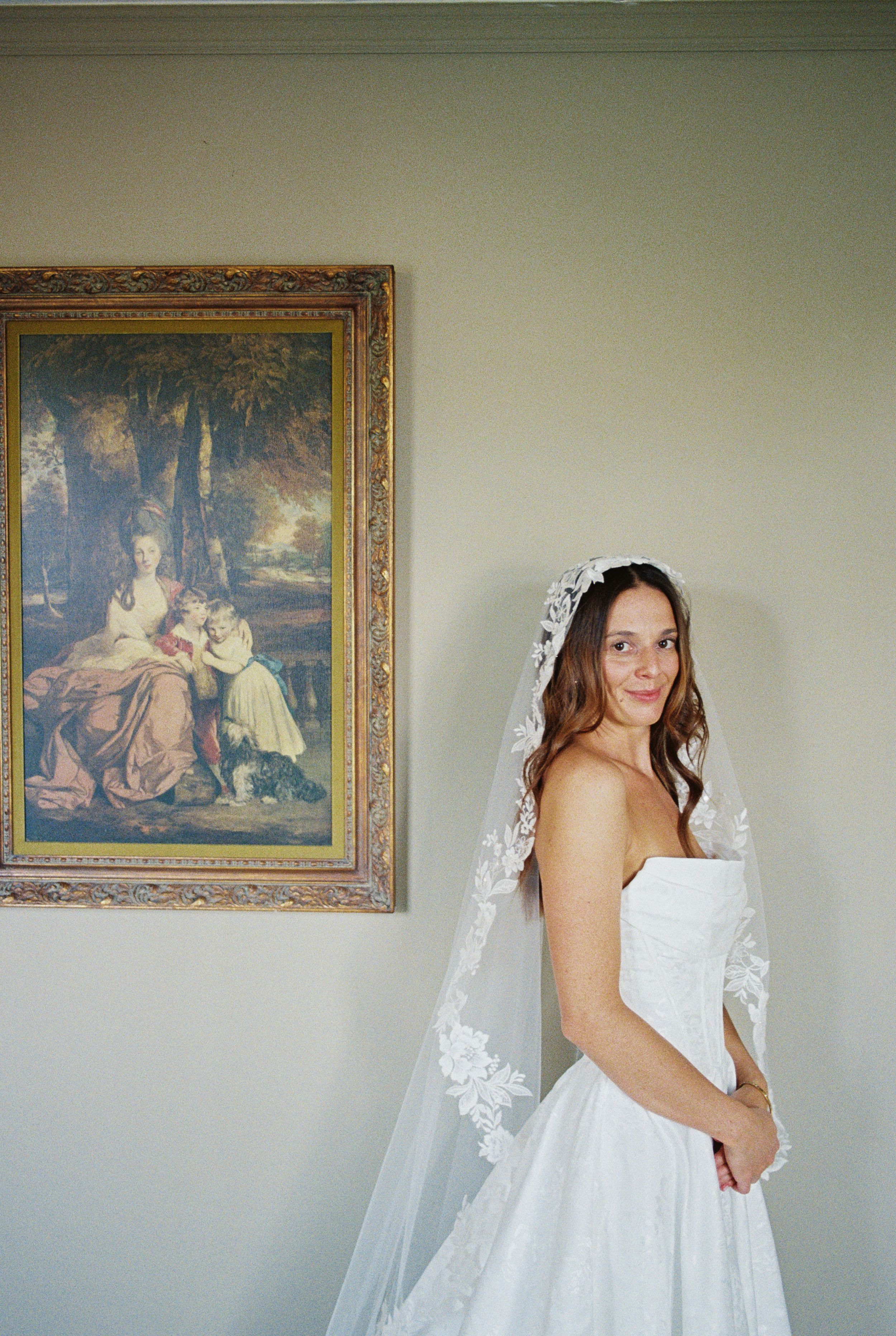 A bride in a white wedding dress and lace veil stands beside an ornate vintage painting in a gilded frame.