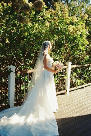 A stunning white wedding gown with flowing train and veil set against lush greenery on a wooden deck in sunlight.