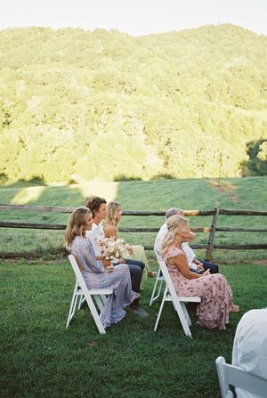 A small group sits on white folding chairs on a grassy lawn with rolling hills in the background during golden hour.