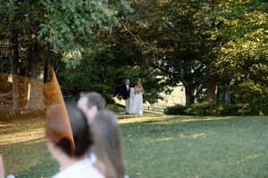A distant couple in wedding attire stands in a garden setting framed through blurred foliage.