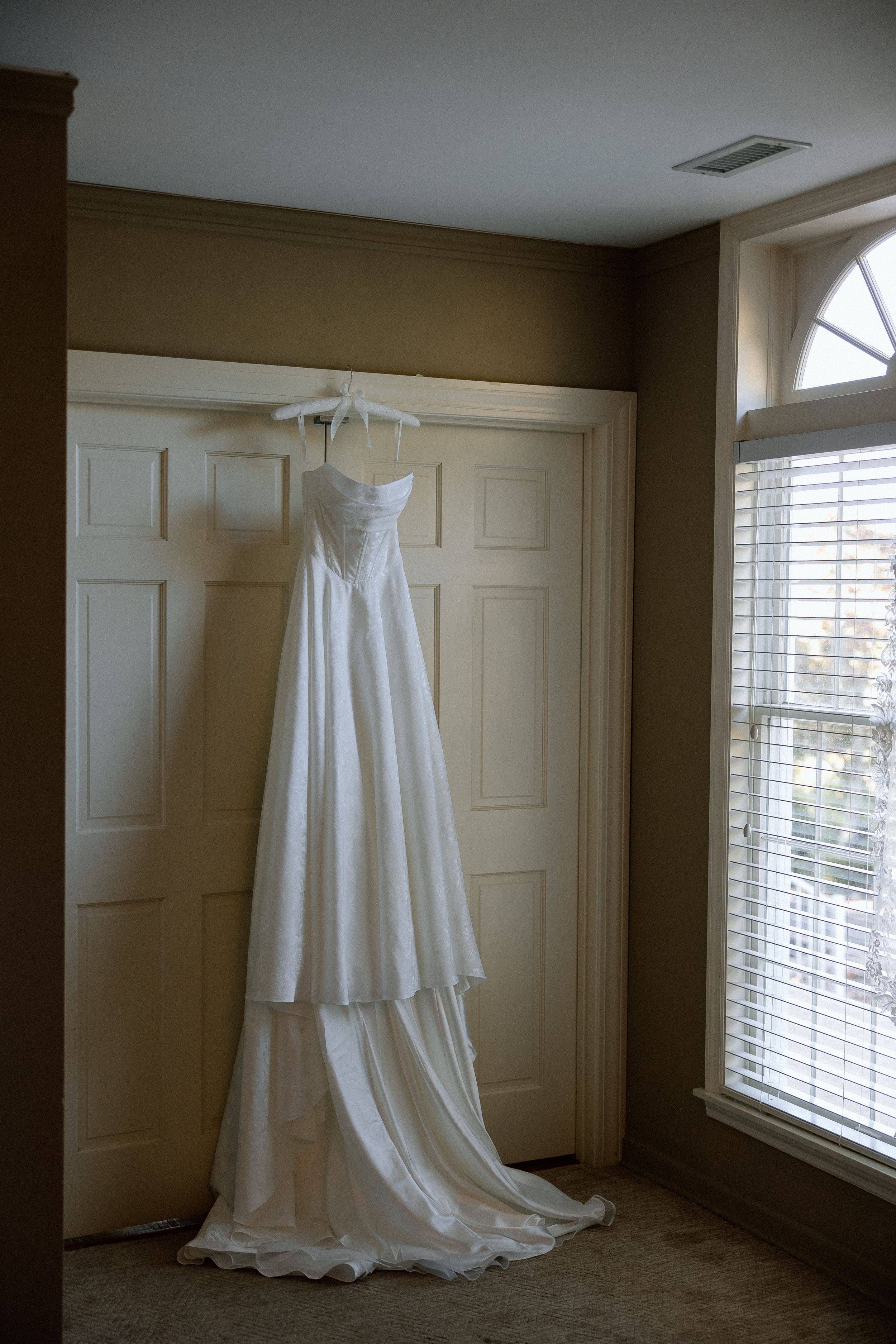 White wedding dress hanging elegantly in a sunlit room with cream walls and window blinds.