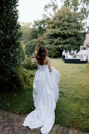A person in a flowing white wedding dress walks along a brick path in a garden setting with guests visible in background.