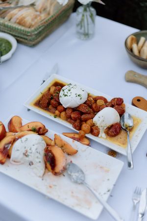 Close-up of burrata cheese plates with roasted tomatoes and peaches on a white tablecloth setting.
