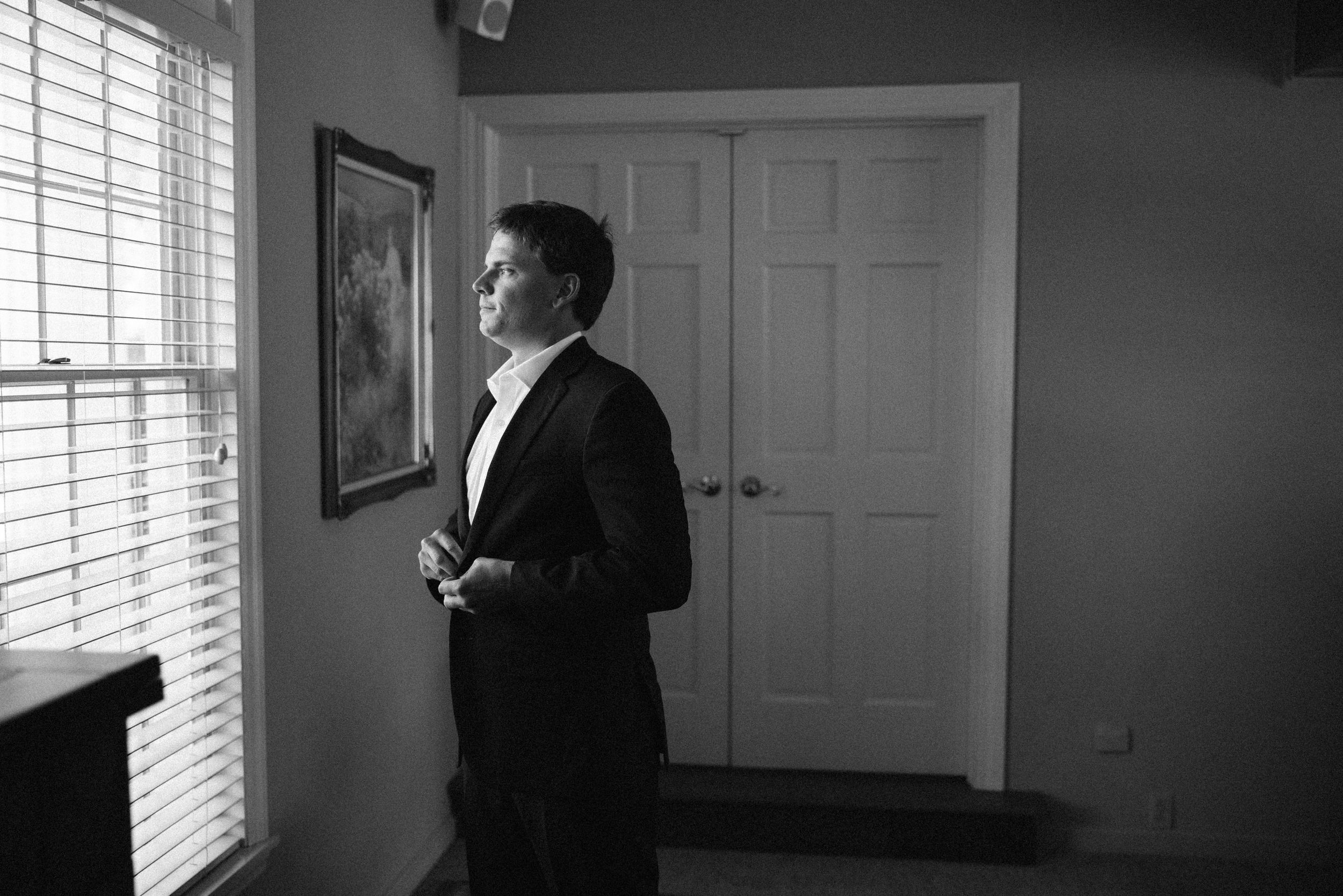 A black and white photograph of a figure standing by a window in formal attire, looking to the side.
