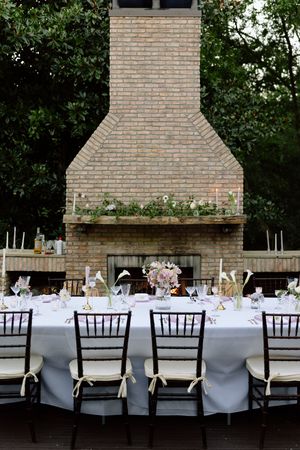 Elegant outdoor wedding reception table setting with white linens and dark chairs in front of a large brick fireplace.