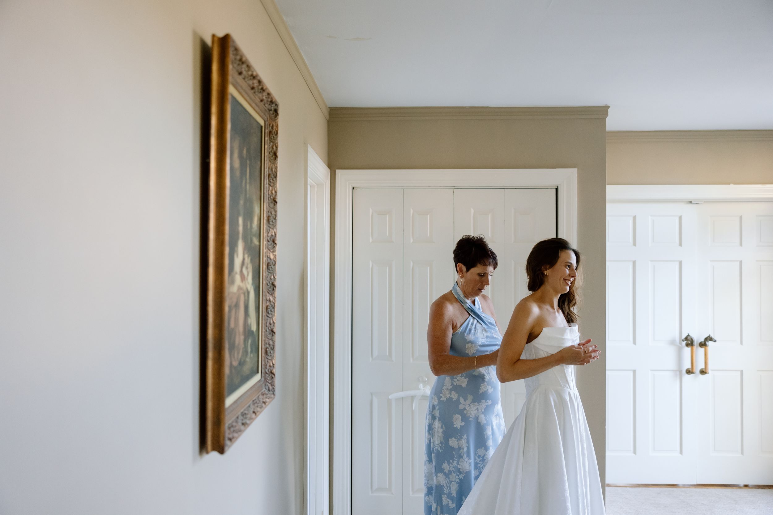 Two people in an elegant room getting ready for a special occasion with one helping adjust the other's white dress.