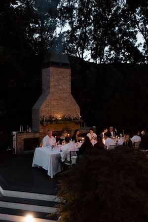 Evening wedding reception setup with candlelit tables and brick fireplace in outdoor setting.