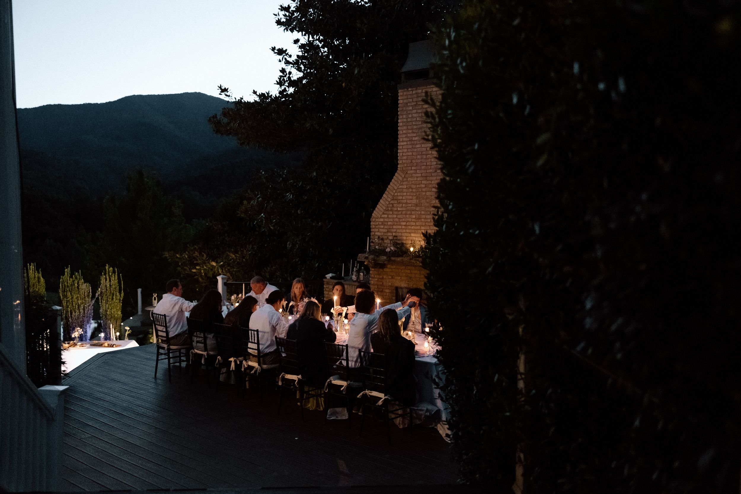 Outdoor dinner party at dusk with guests seated at a long table on a deck overlooking mountain scenery.