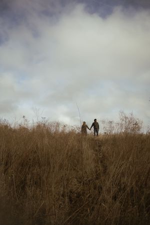 Couple walking through tall brown grass under dramatic cloudy skies.
