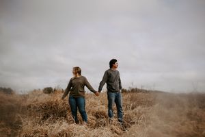 A couple holds hands while standing in a field of golden grass under cloudy skies.