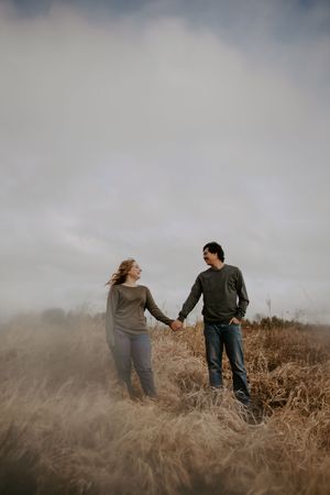 Two people walk hand in hand through tall grass on a moody, overcast day.