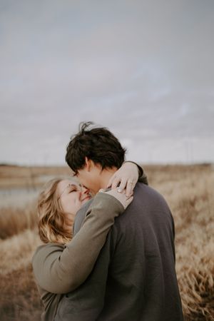 A romantic couple embraces in a field under moody skies wearing neutral sweaters.