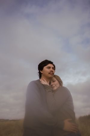 A couple shares an affectionate embrace against a dramatic cloudy sky backdrop.
