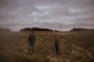 A moody landscape photograph of figures walking through a golden field under overcast skies.