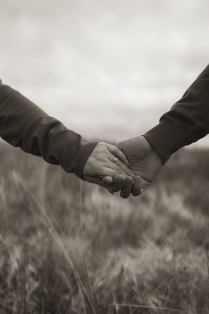 Grayscale image of joined hands floating above wispy grass with moody clouds overhead.
