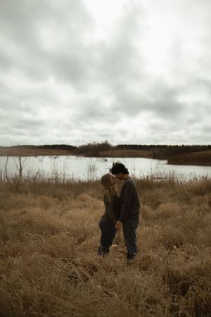 A couple embraces in a field of tall grass under moody cloudy skies.