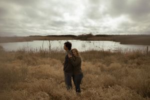 A couple stands together overlooking a serene lake on an overcast day in a grassy field.